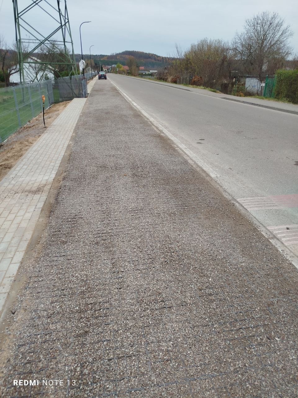 Przygotowanie podłoża pod chodnik z kostki: wyrównany teren z siatką wzmacniającą, obok jezdnia i chodnik z kostki. Słup wysokiego napięcia w tle.