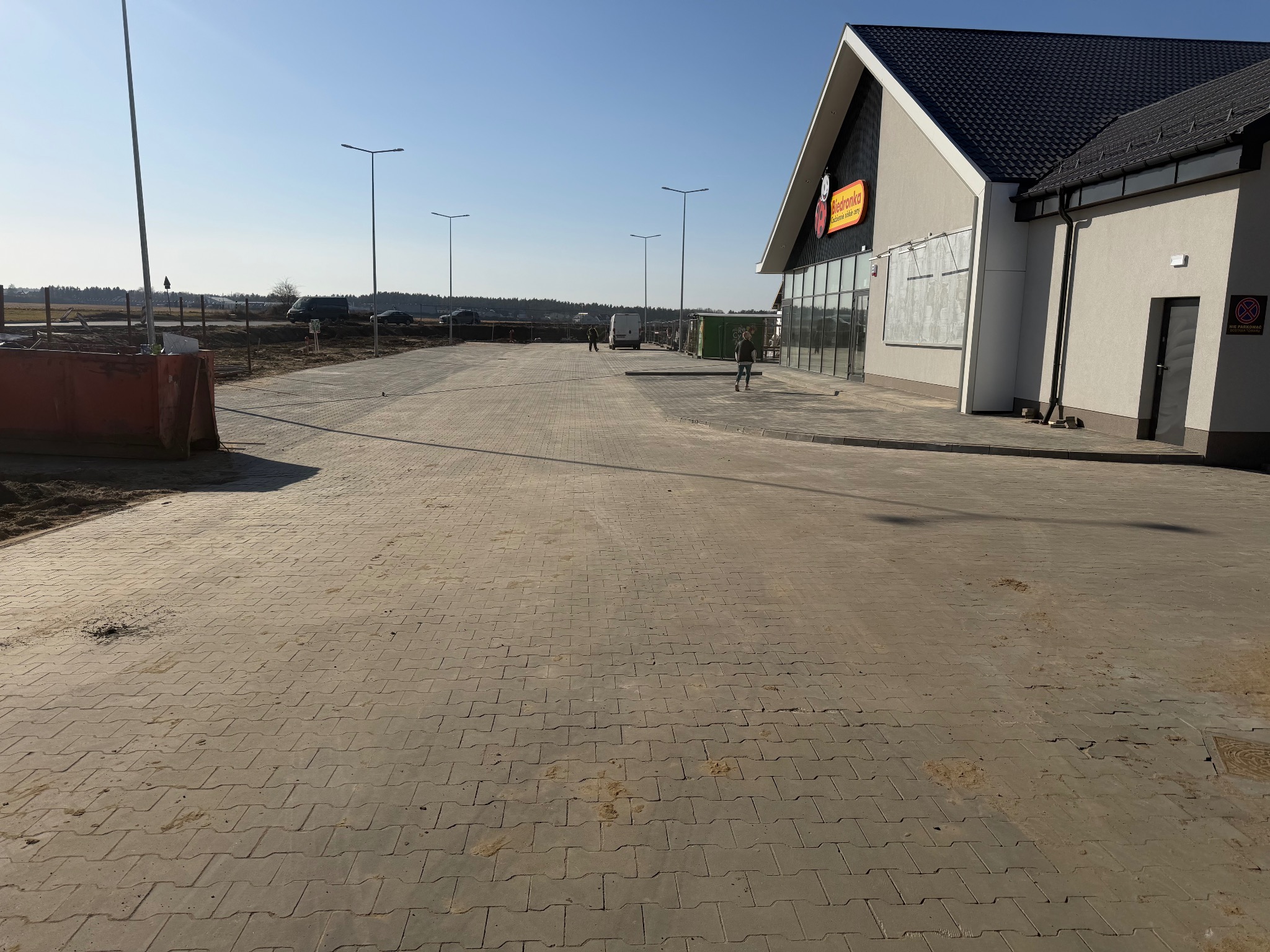 Rozległy parking z kostki brukowej przed budynkiem sklepu, oświetlony słupami. W tle widoczne prace ziemne i zaparkowane pojazdy.