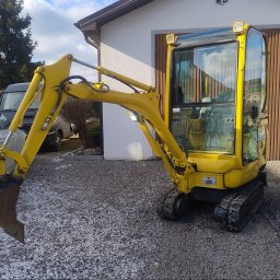 STELP - Żółta mini koparka na żwirowym podłożu, z widocznym budynkiem w tle. Koparka z łyżką skierowaną w lewo, oświetlenie robocze na kabinie i ramieniu.