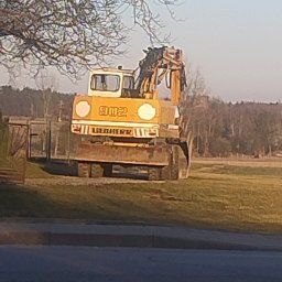 STELP - Żółta koparka Liebherr 902 na trawiastym terenie, widok z przodu, drzewo z lewej strony kadru. W tle las i błękitne niebo.
