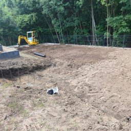 STELP - Żółta koparka na placu budowy, przygotowany teren z widocznymi elementami fundamentów i zielenią w tle. Prace ziemne w toku.