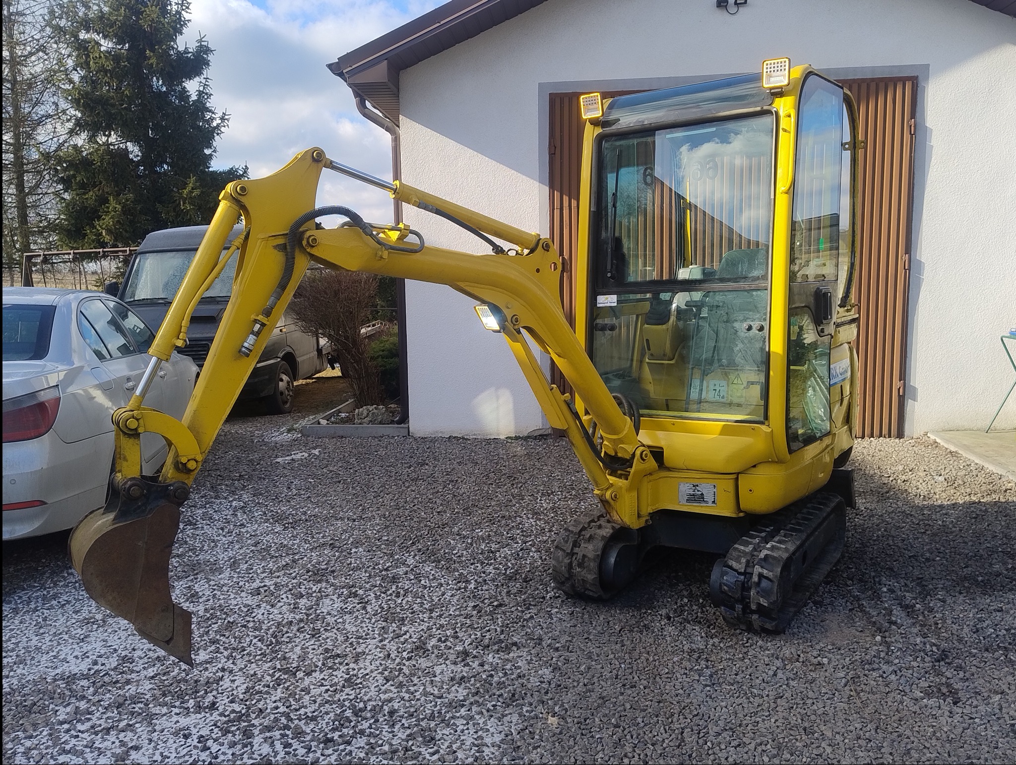 Żółta mini koparka na żwirowym podłożu, z widocznym budynkiem w tle. Koparka z łyżką skierowaną w lewo, oświetlenie robocze na kabinie i ramieniu.