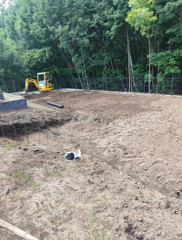 Żółta koparka na placu budowy, przygotowany teren z widocznymi elementami fundamentów i zielenią w tle. Prace ziemne w toku.