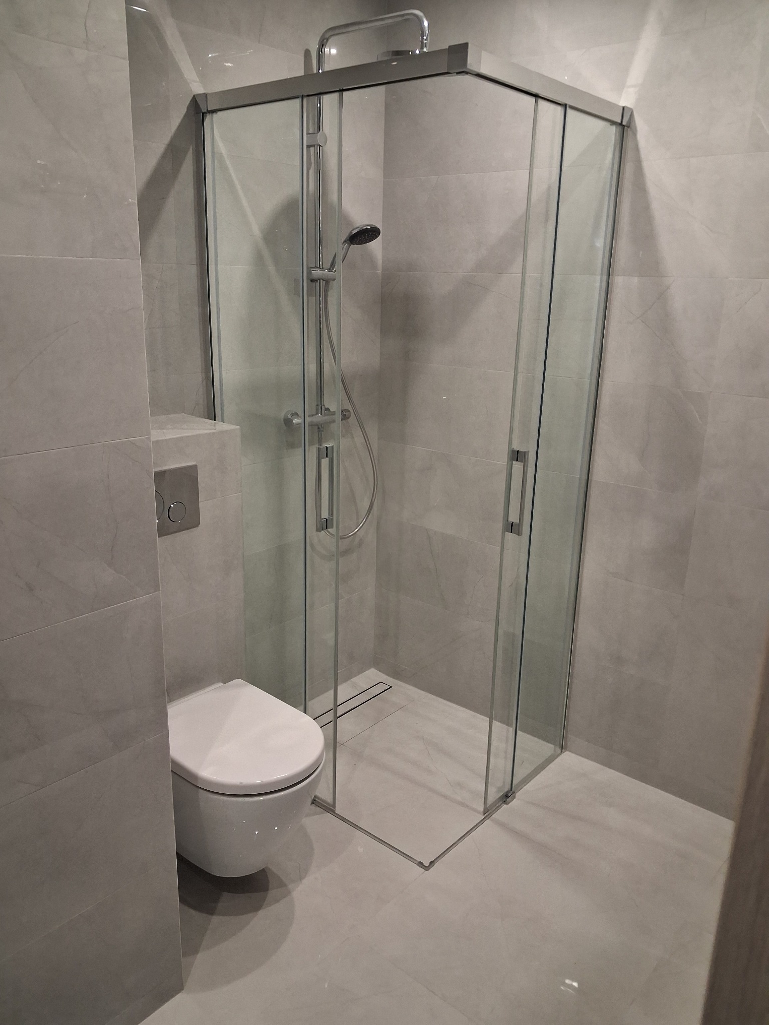 Nowoczesna łazienka w szarościach: kabina prysznicowa z transparentnego szkła, podwieszana toaleta i płytki imitujące marmur. Minimalistyczny design.