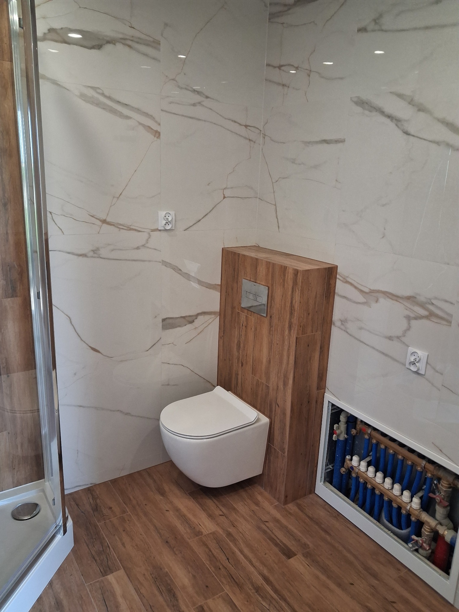 Wykończona łazienka z podwieszaną toaletą, płytkami imitującymi marmur i drewno. Widoczna instalacja ogrzewania podłogowego w szafce. Kabina prysznicowa z elementami chromowanymi.