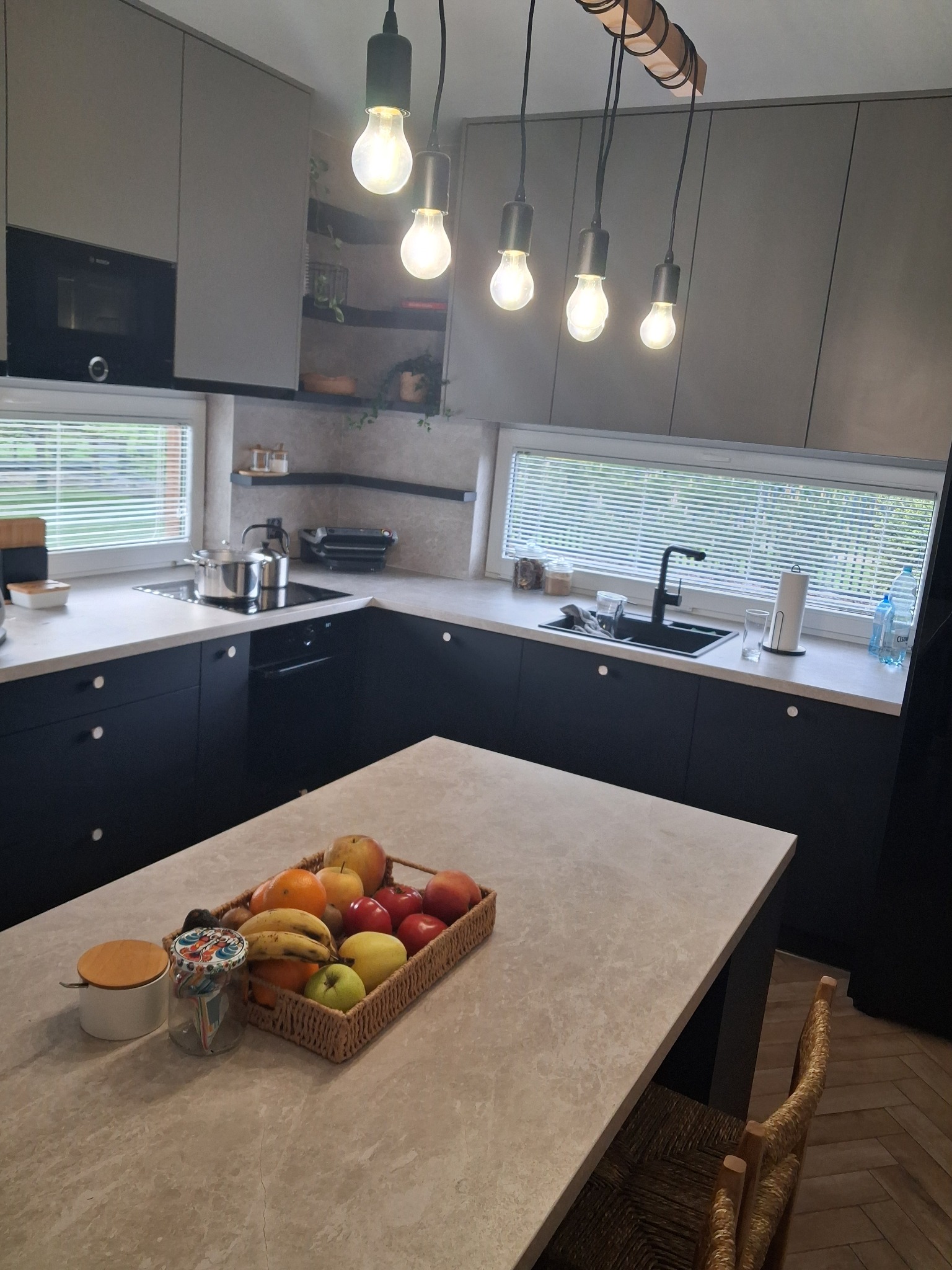Nowoczesna kuchnia z wyspą i owocami. Czarne szafki, szare fronty górne, drewniane akcenty oświetlenia. Minimalistyczny design, jasne blaty, rolety w oknach.