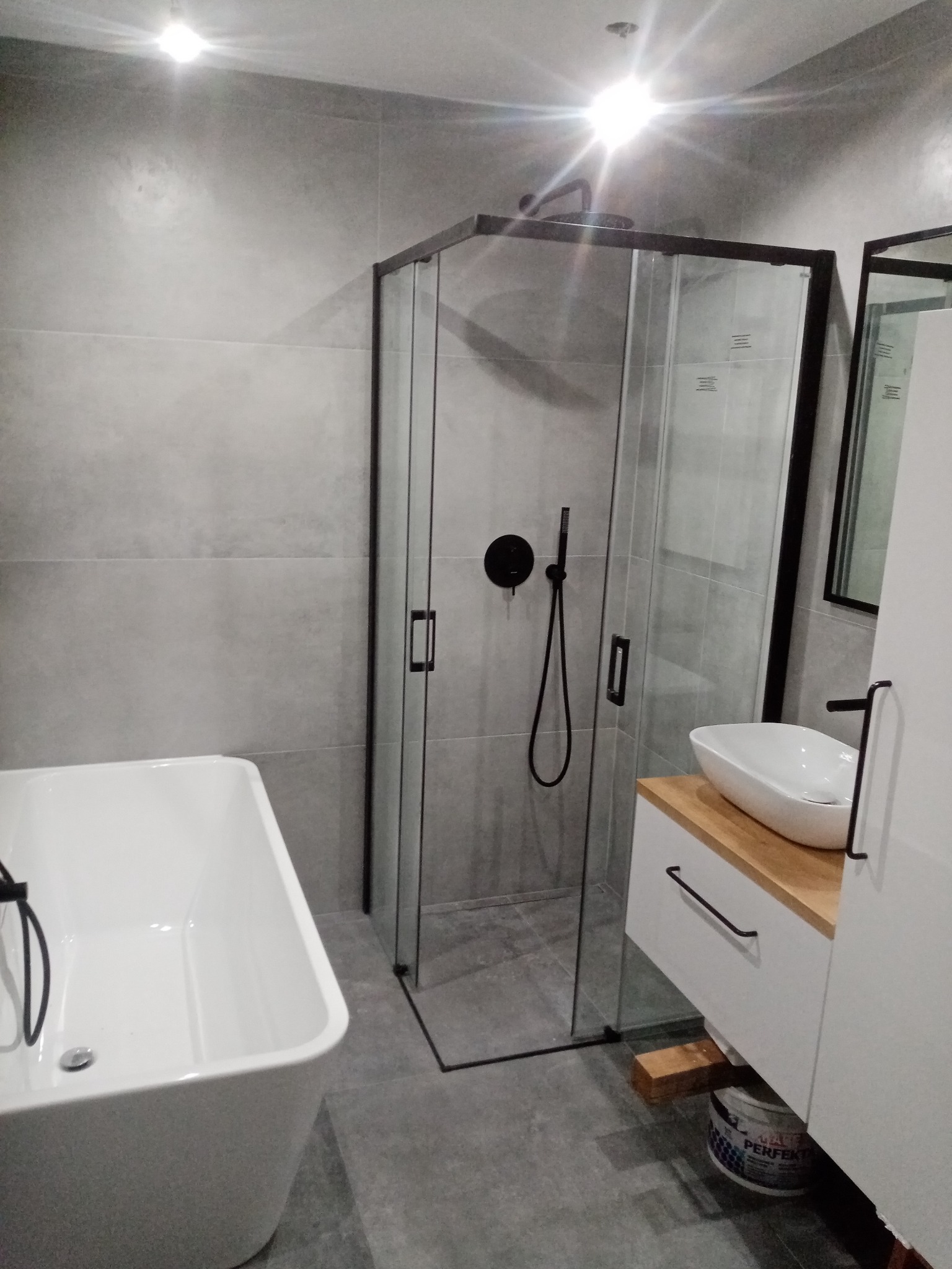 Nowoczesna łazienka w szarościach: kabina prysznicowa z czarnymi detalami, wolnostojąca wanna i umywalka nablatowa na szafce z drewnianym blatem.