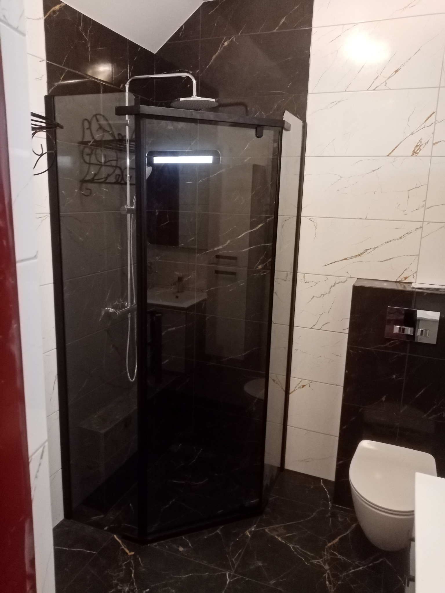 Nowoczesna kabina prysznicowa z czarną ramą i deszczownicą, w łazience wykończonej płytkami imitującymi marmur w odcieniach bieli i czerni. Widoczna toaleta i umywalka.