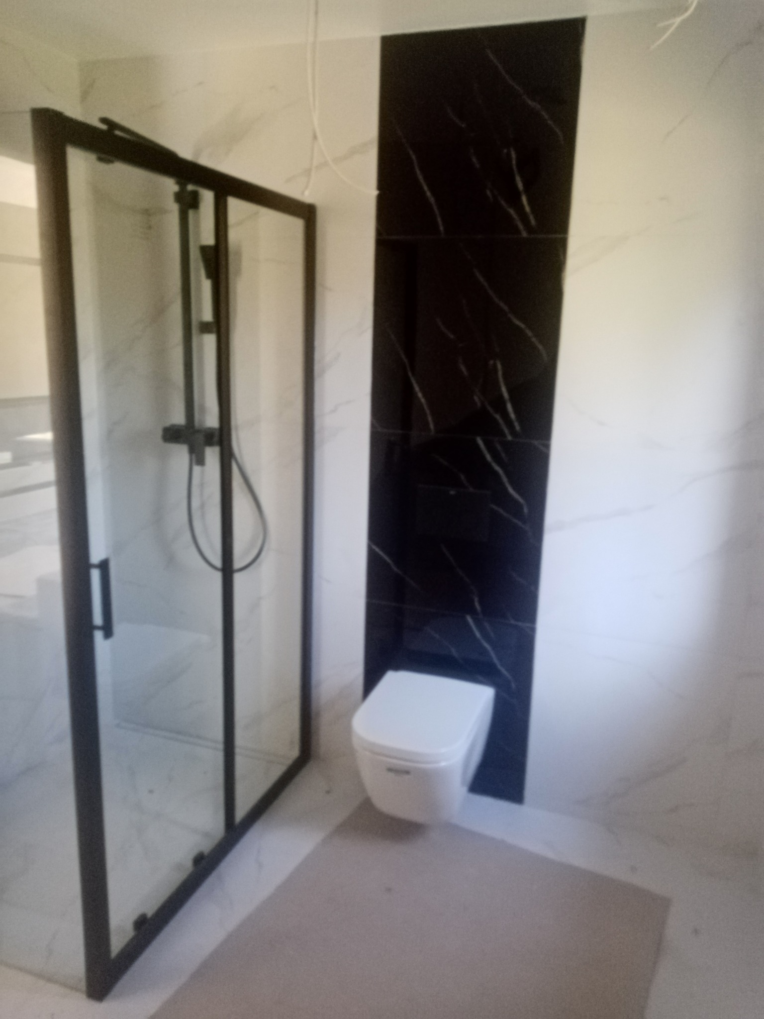 Nowoczesna łazienka po remoncie: kabina prysznicowa z czarną ramą, biała toaleta i marmurowe płytki w odcieniach bieli i czerni. Minimalistyczny design.