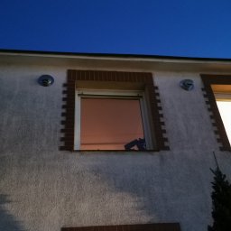 DIAMCORE - Elewacja budynku z widocznymi dwoma okrągłymi wlotami rekuperacji, umieszczonymi symetrycznie nad oknami. Tynk strukturalny, ceglana obramówka okien.