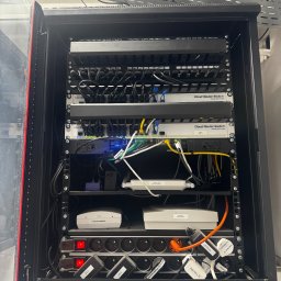 Grupa Promes sp. z o.o. - Szafa rack z zamontowanymi urządzeniami sieciowymi: switche, routery, patch panel. Widoczne okablowanie i zasilacze. Otwarta szafa z czerwonym akcentem na krawędzi.