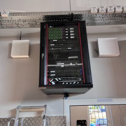 Grupa Promes sp. z o.o. - Zamontowana szafa rack z urządzeniami sieciowymi i zasilającymi, widoczne kable i skrzynki zasilające na ścianie. Na dole drabina i fragment pomieszczenia biurowego.