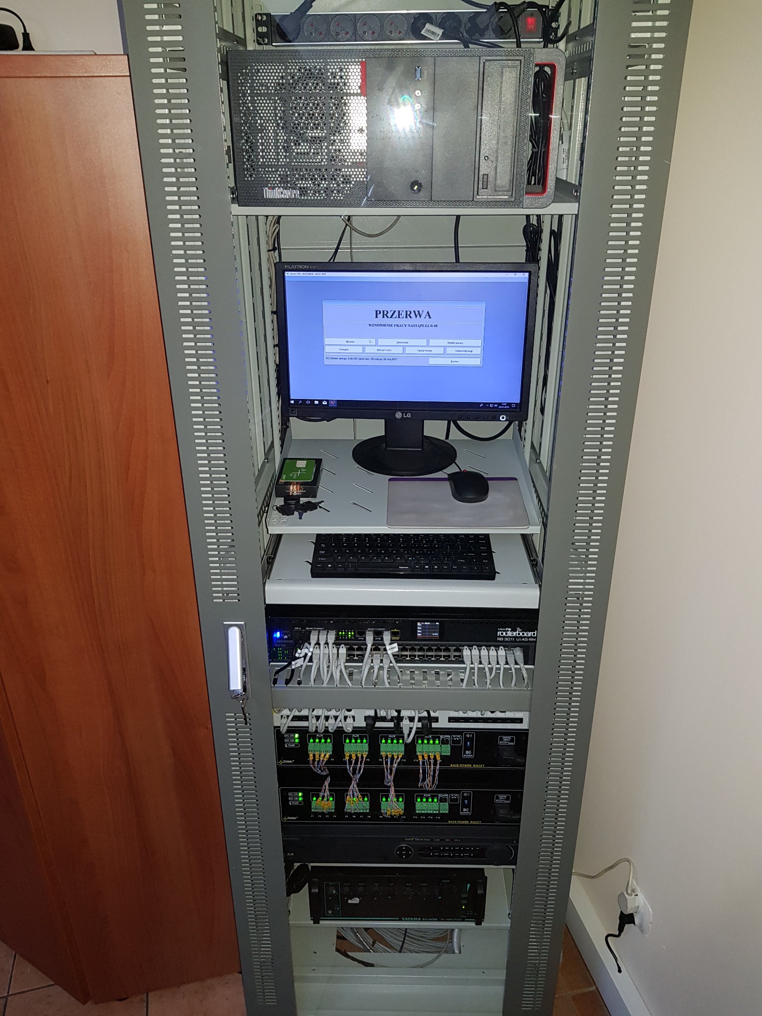 Szafa rack z serwerem, monitorem, klawiaturą, switchem i zasilaczami. Widoczne kable i wtyczki. Sprzęt sieciowy w szarej obudowie. Monitor LG z komunikatem o przerwie.