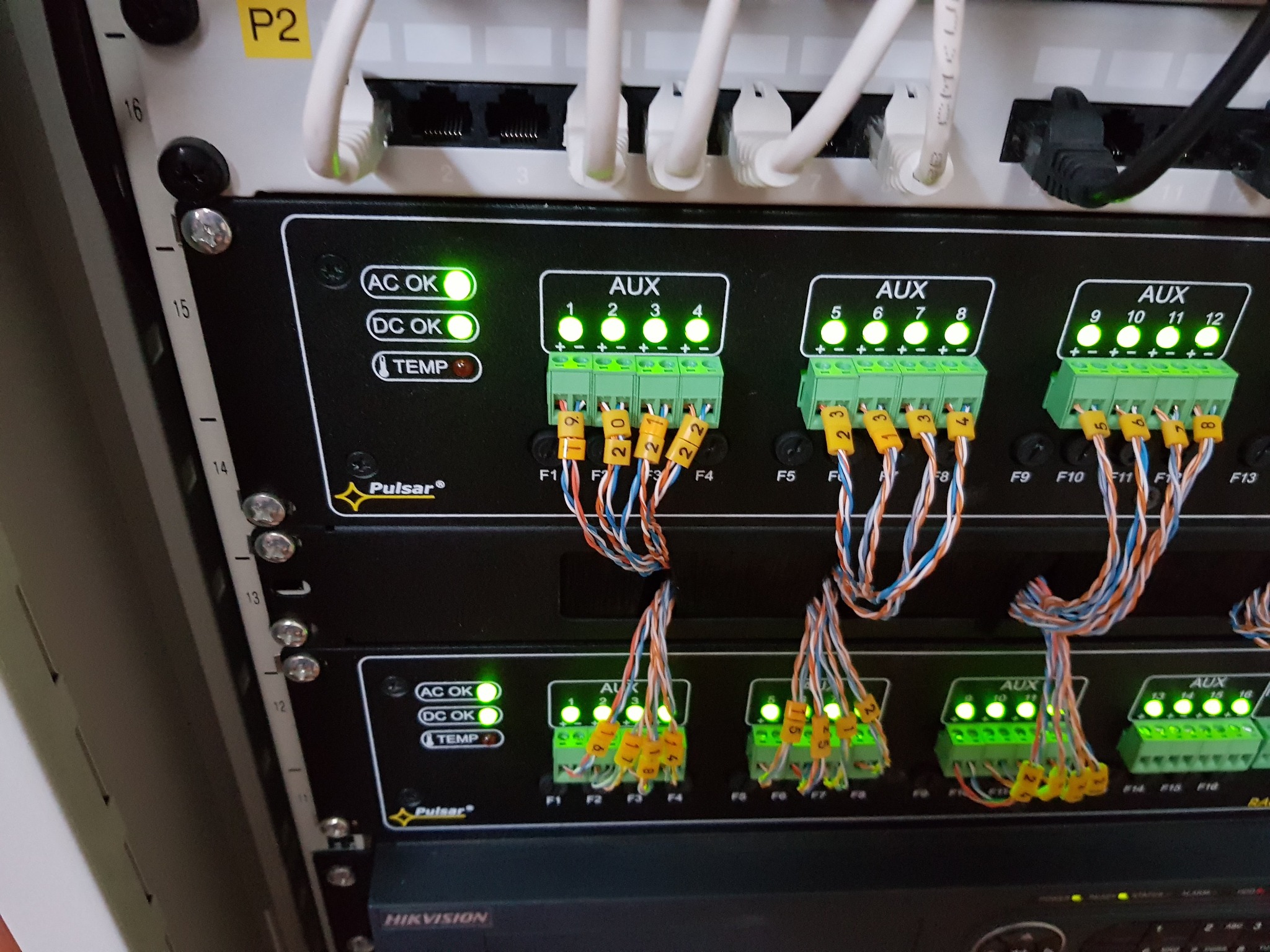 Panel sterowania z podłączonymi kablami i diodami LED sygnalizującymi status. Widoczne oznaczenia AUX, AC OK, DC OK, TEMP oraz logo Pulsar i Hikvision.