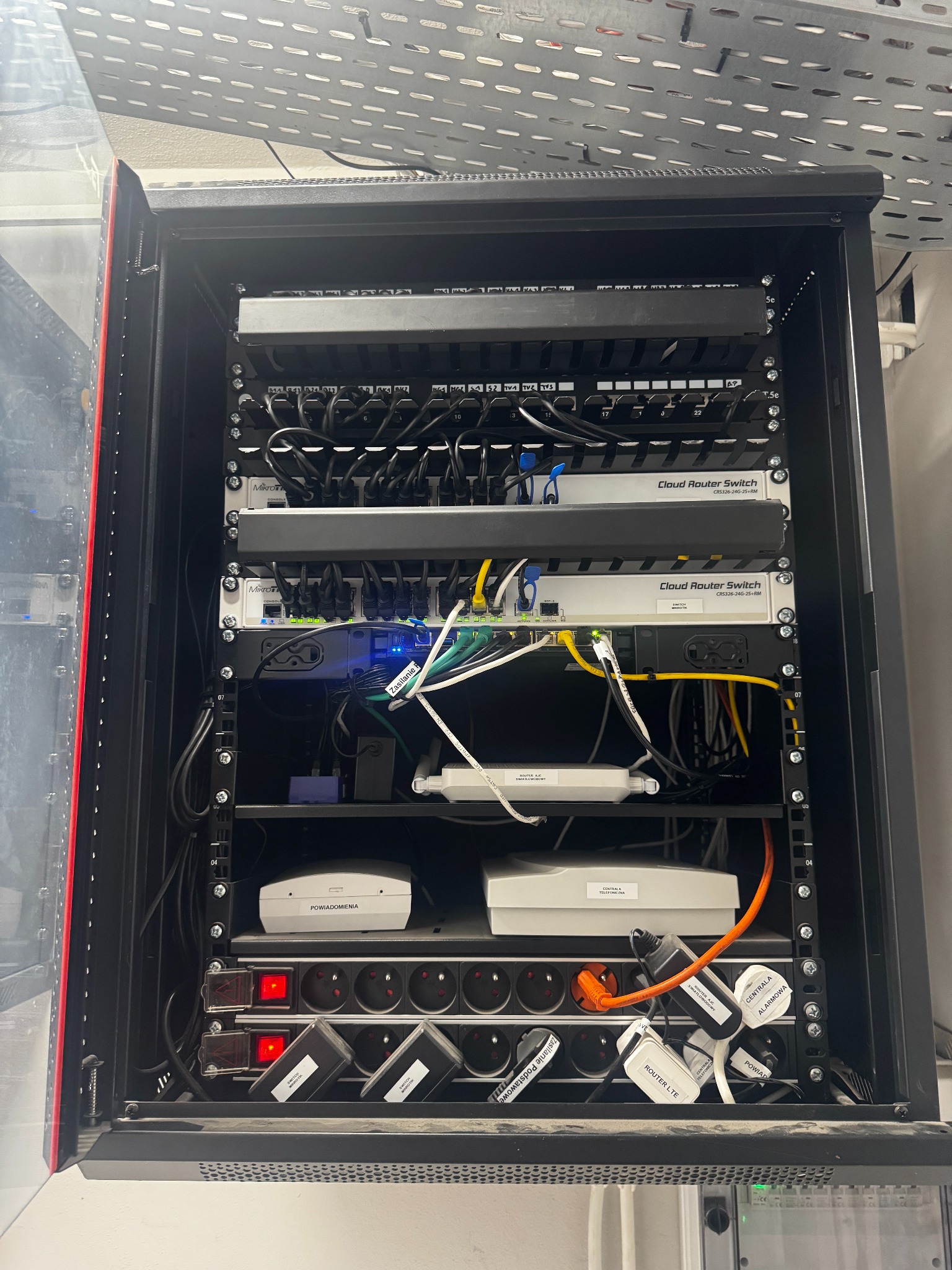 Szafa rack z zamontowanymi urządzeniami sieciowymi: switche, routery, patch panel. Widoczne okablowanie i zasilacze. Otwarta szafa z czerwonym akcentem na krawędzi.