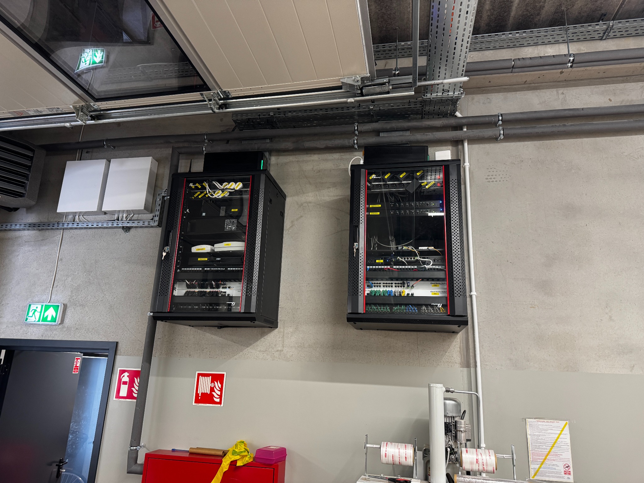 Dwie szafy rack z zainstalowanymi urządzeniami sieciowymi, zamontowane na ścianie w pomieszczeniu technicznym. Widoczne kable, switche i inne elementy infrastruktury IT.