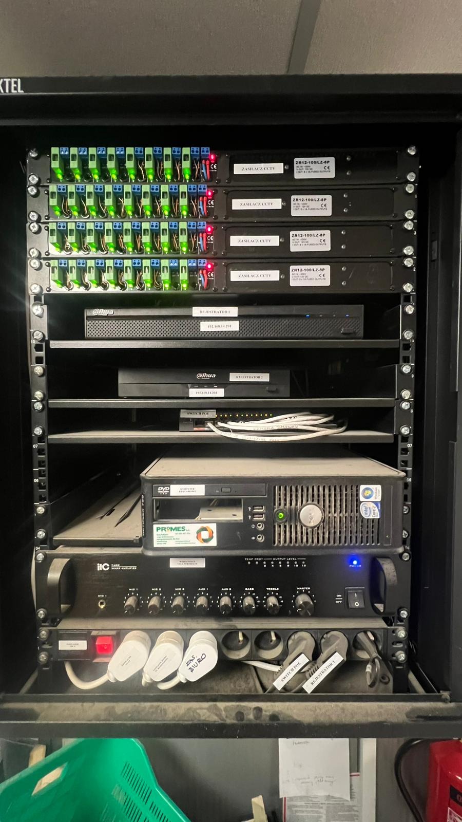 Szafa rack z zasilaczami CCTV, rejestratorami Dahua, switchem PoE i wzmacniaczem ITC. Widoczne kable i oznaczenia. Sprzęt zakurzony, co sugeruje długotrwałą eksploatację.