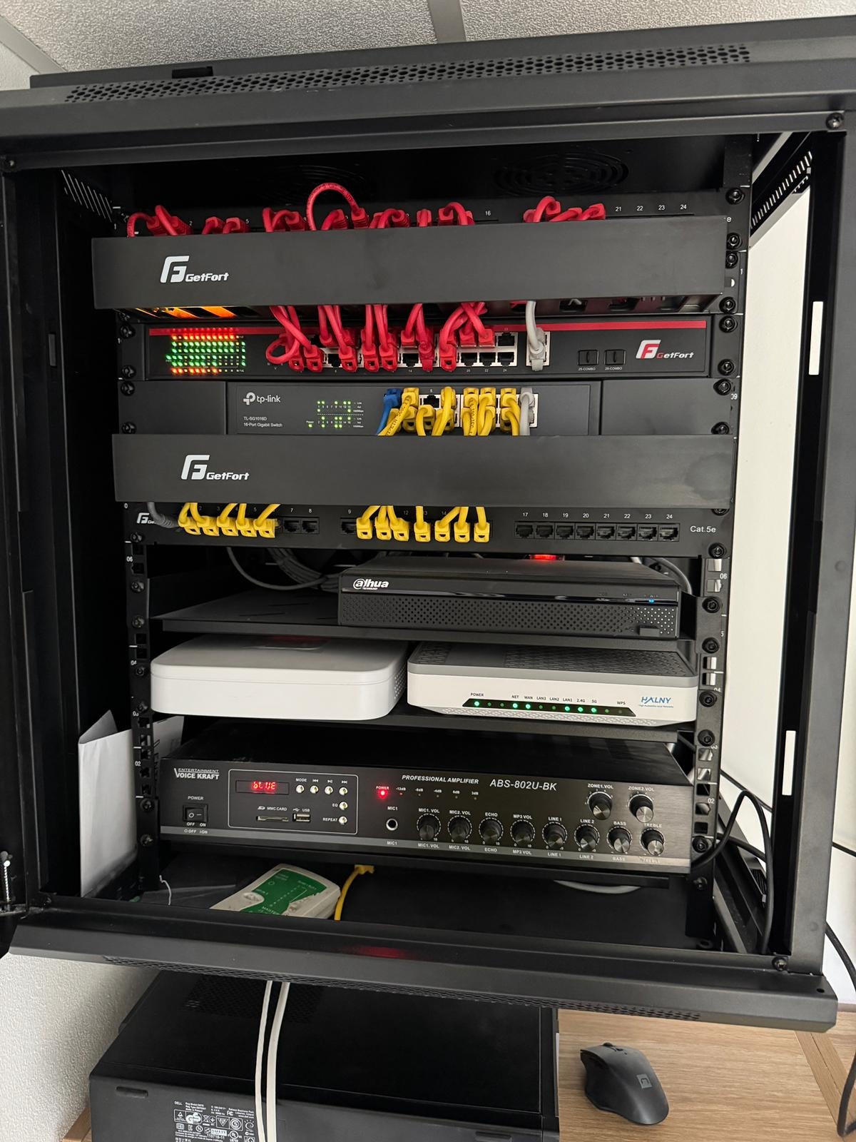Otwarta szafa rack z zainstalowanymi urządzeniami sieciowymi: routery, switche, rejestrator Dahua, wzmacniacz Voice Kraft i okablowanie. Widoczne porty Ethernet i diody LED.