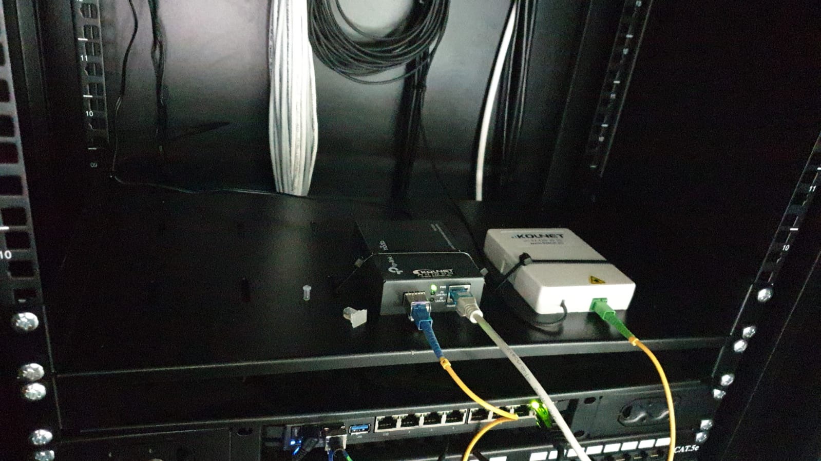 Zamknięta szafa rack z podłączonymi urządzeniami sieciowymi Kounet i TP-Link, widoczne kable światłowodowe i ethernetowe. Elementy instalacji sieci komputerowej.