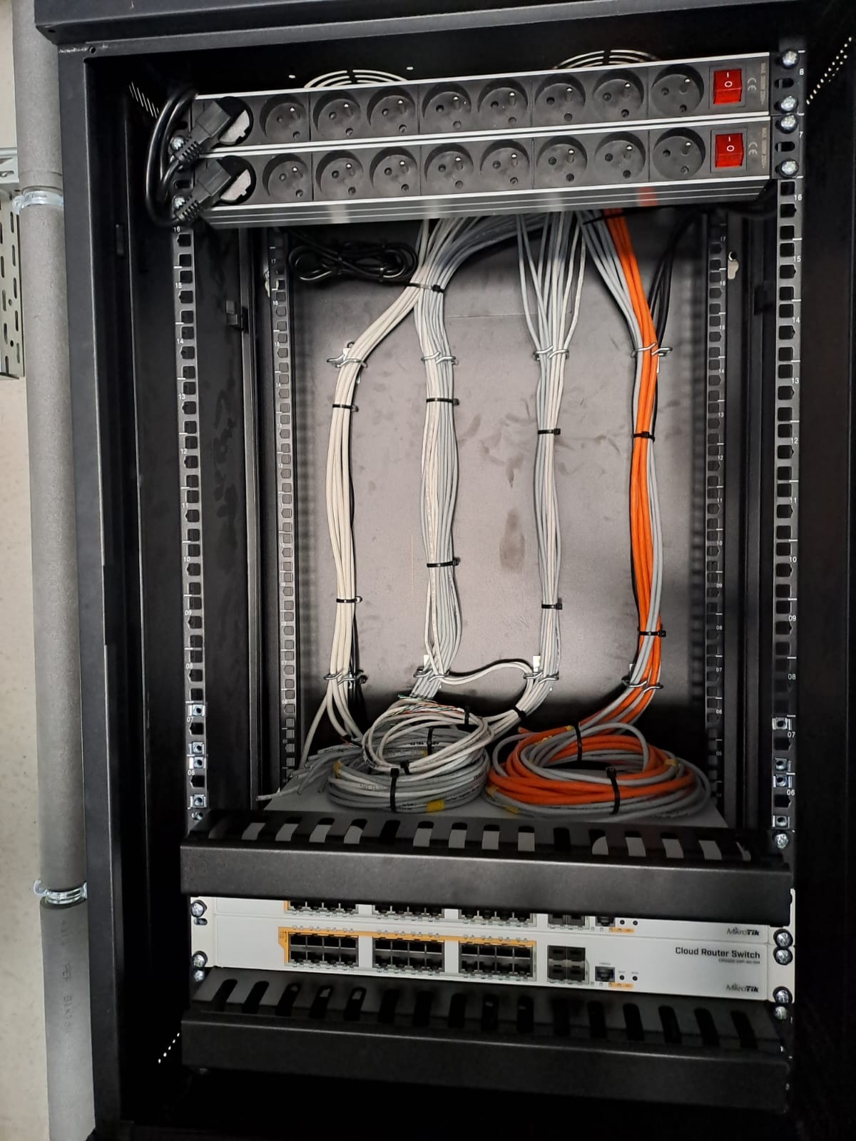 Szafa rack z uporządkowanymi kablami sieciowymi (szare i pomarańczowe) i listwą zasilającą. Widoczny switch MikroTik Cloud Router Switch. Estetyczna instalacja.
