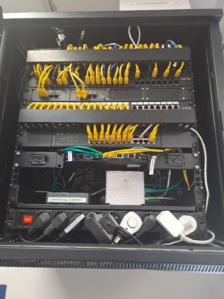 Szafa rack z patch panelem, switchami i okablowaniem strukturalnym. Żółte kable patch cord CAT6 i CAT5e. Widoczny router ZTE i listwa zasilająca z włączonym zasilaniem.