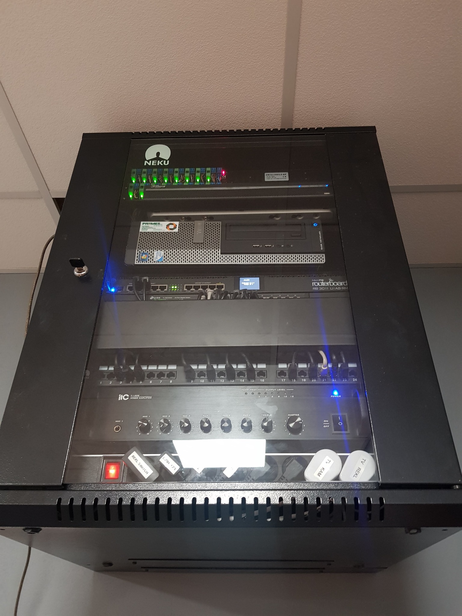 Zamknięta szafa rack z zainstalowanymi urządzeniami sieciowymi i wzmacniaczem ITC. Widoczne kable, diody LED i etykiety. Sprzęt NEKU, Promes i Routerboard.