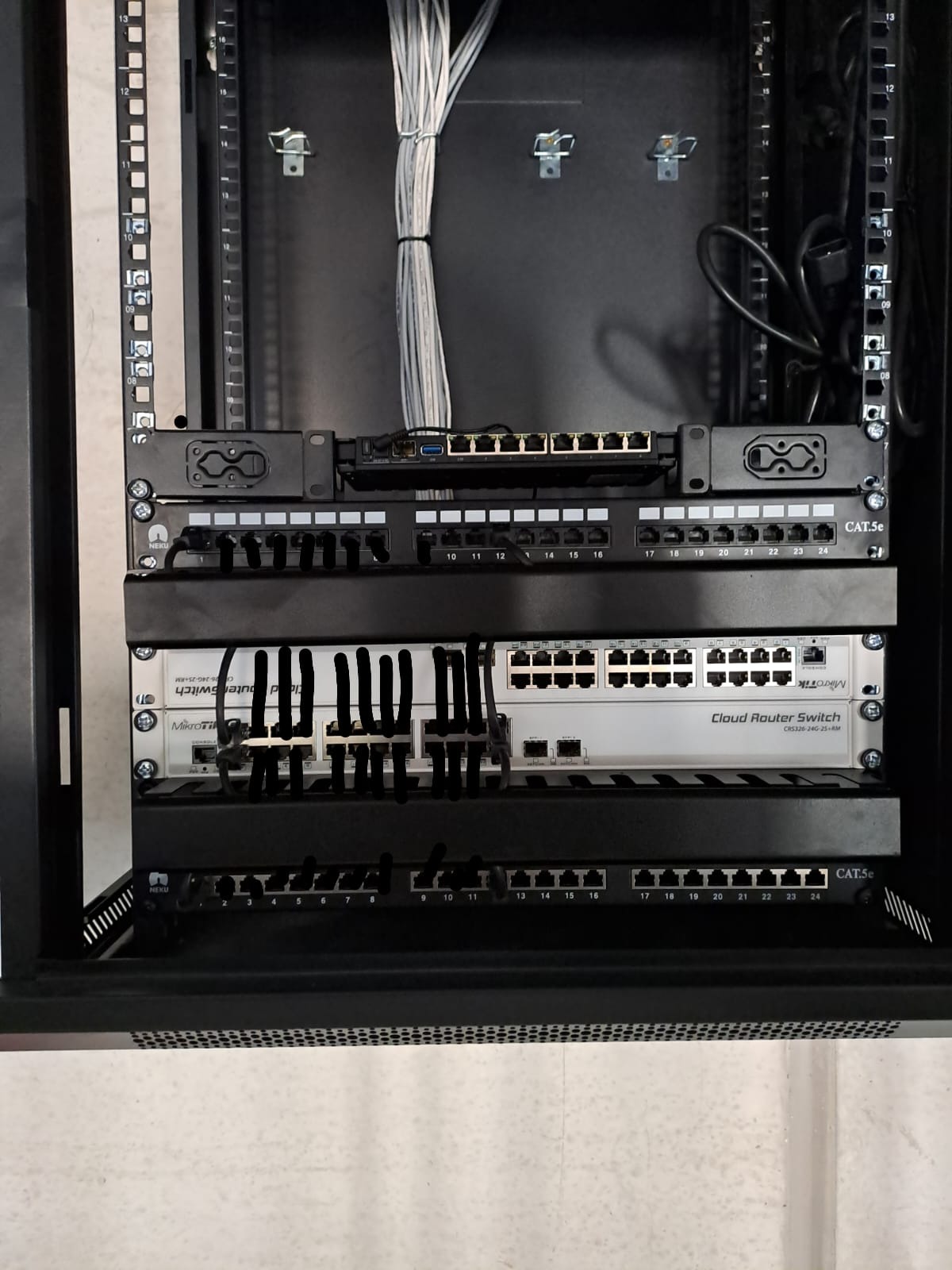 Zamknięta szafa rack z zamontowanymi panelami krosowniczymi, switchem Mikrotik Cloud Router Switch i okablowaniem. Widoczne porty RJ45 i oznaczenia CAT.5e.