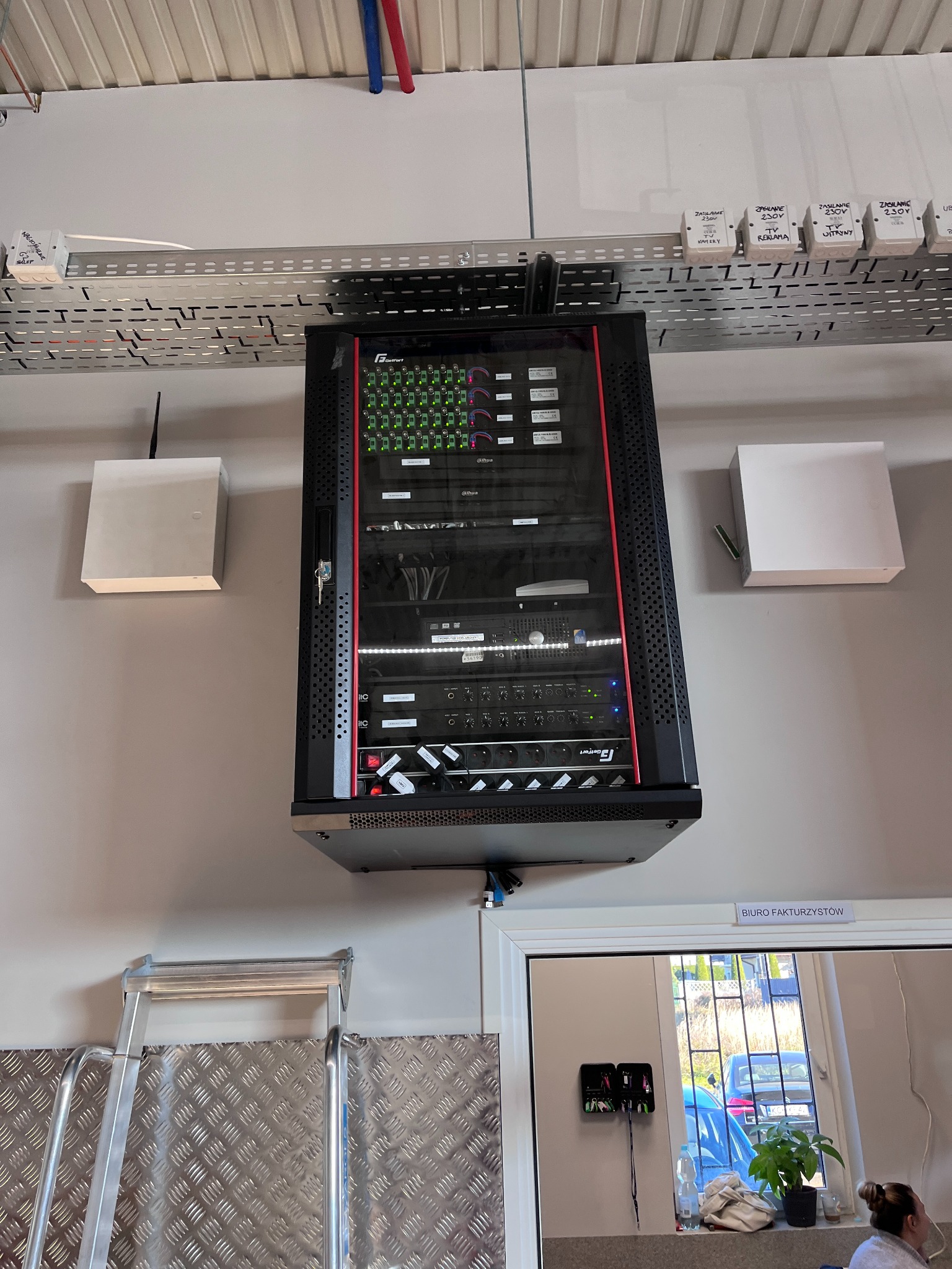 Zamontowana szafa rack z urządzeniami sieciowymi i zasilającymi, widoczne kable i skrzynki zasilające na ścianie. Na dole drabina i fragment pomieszczenia biurowego.