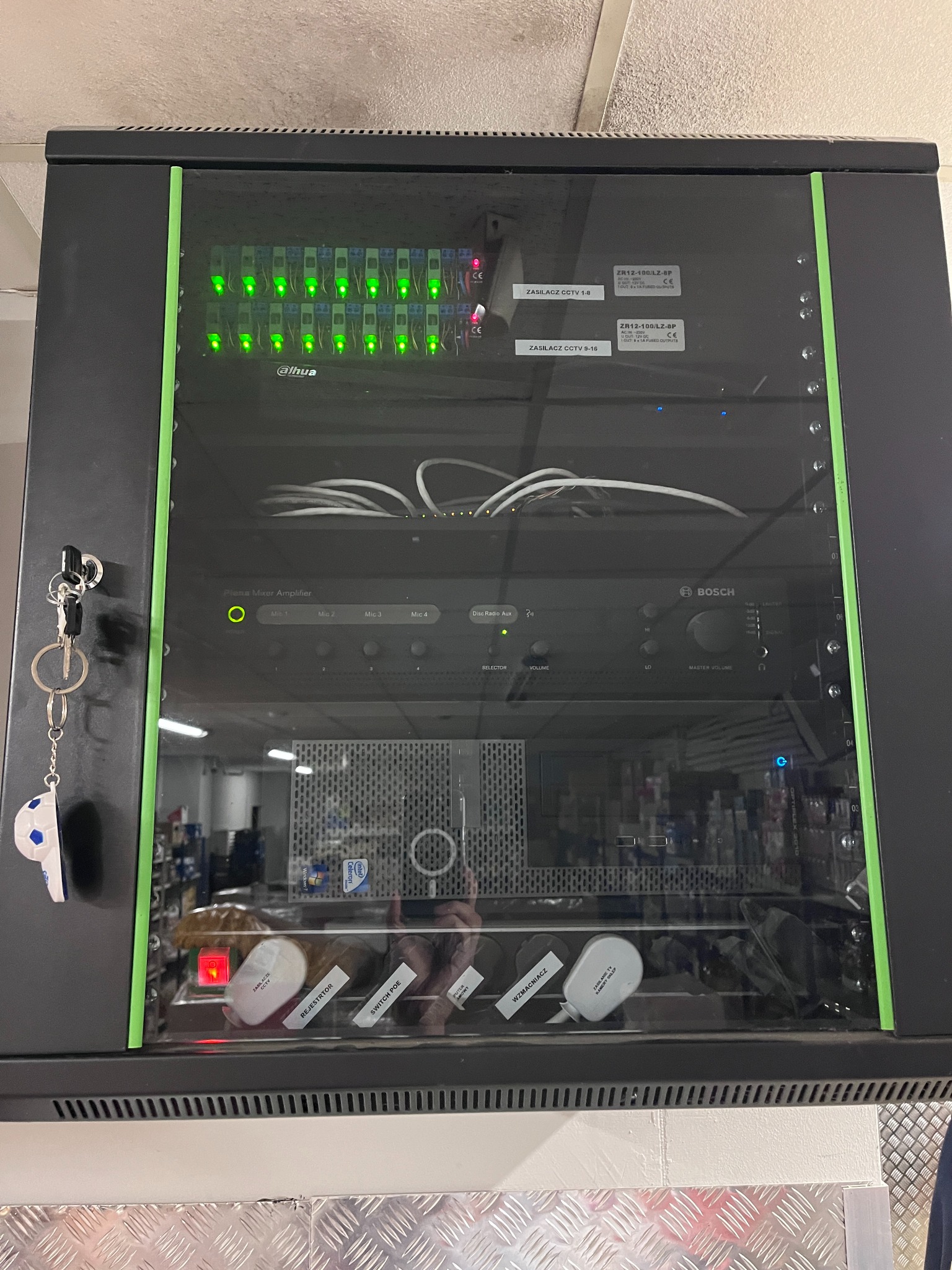 Szafa rack z zainstalowanymi urządzeniami: zasilacze CCTV, wzmacniacz Bosch, rejestrator, switch PoE. Widoczne kable i diody sygnalizacyjne. Klucze wiszą na boku szafy.