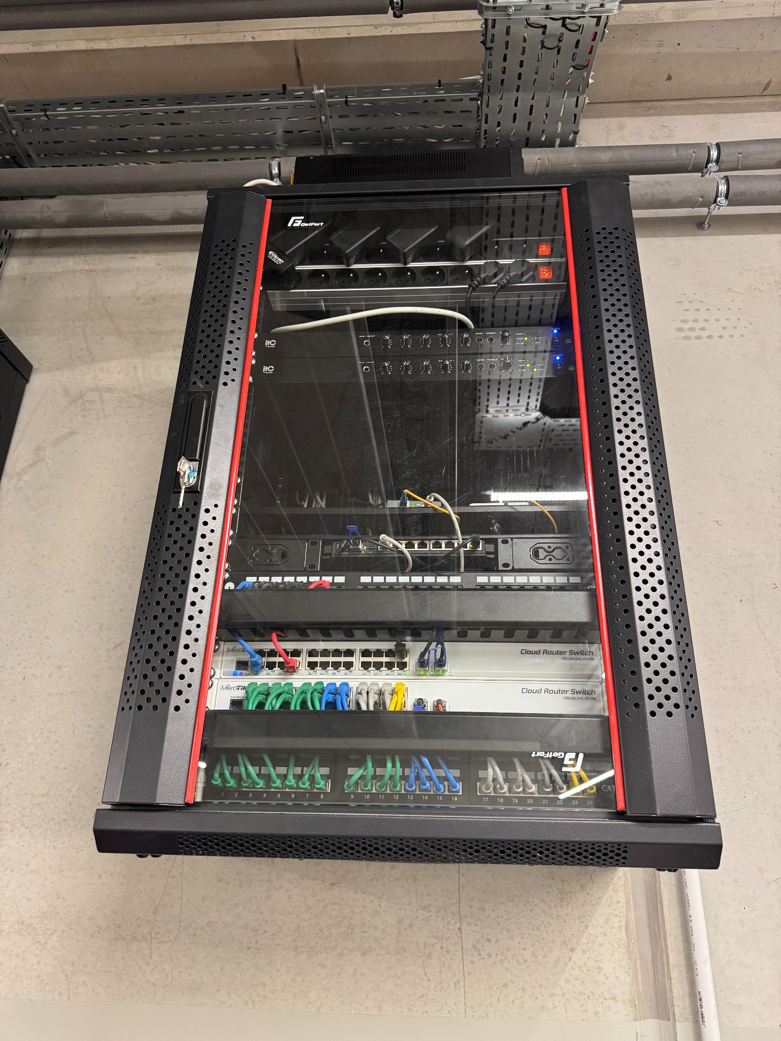 Zamknięta szafa rack z zainstalowanymi urządzeniami sieciowymi, w tym routerami i przełącznikami, z widocznymi kablami. Szafa w kolorze czarnym z czerwonymi akcentami.