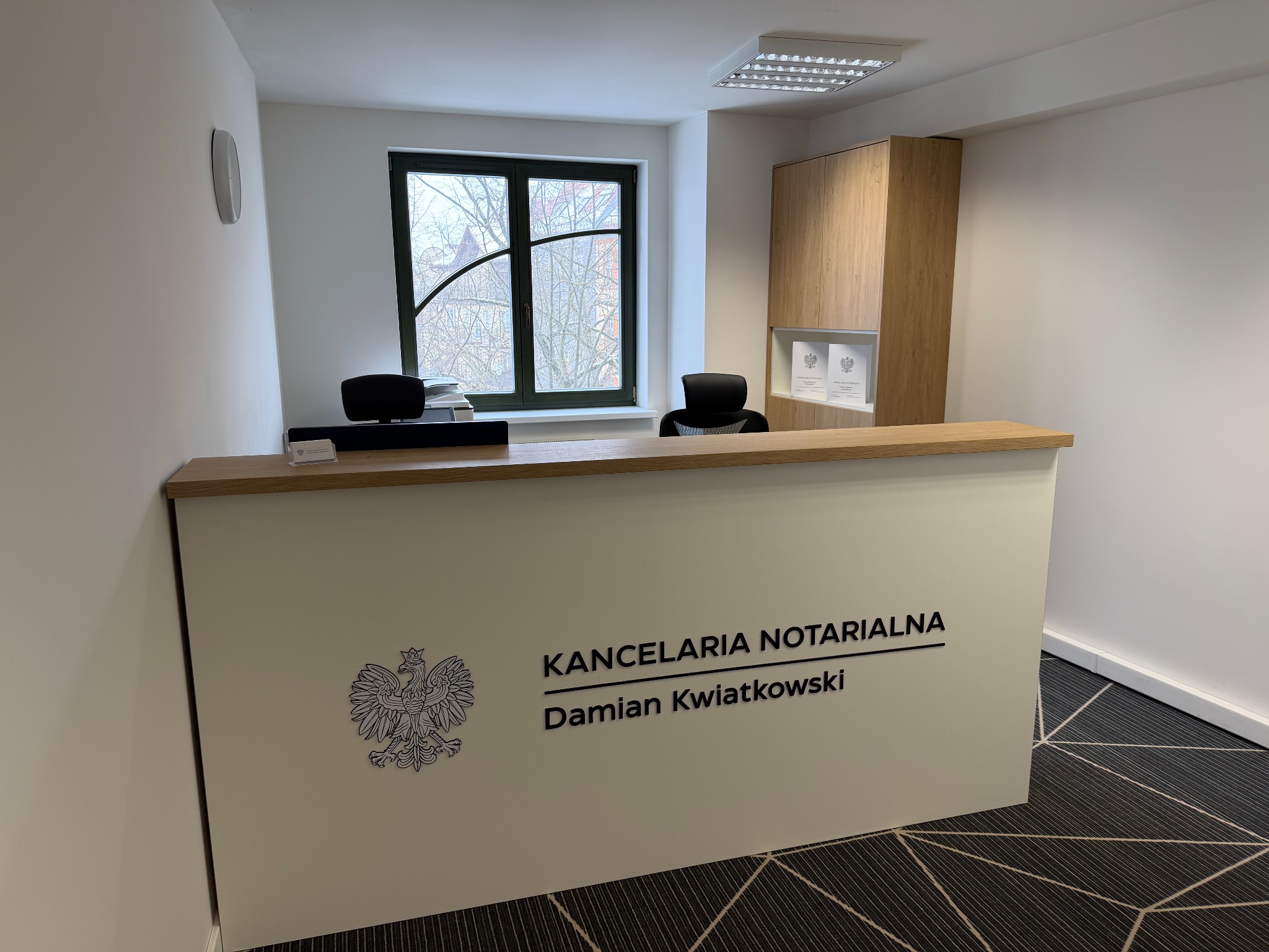 Recepcja Kancelarii Notarialnej Damian Kwiatkowski z godłem Polski, widok z przodu, nowoczesne wnętrze z oknem i drewnianą szafą, czarno-biała wykładzina podłogowa.