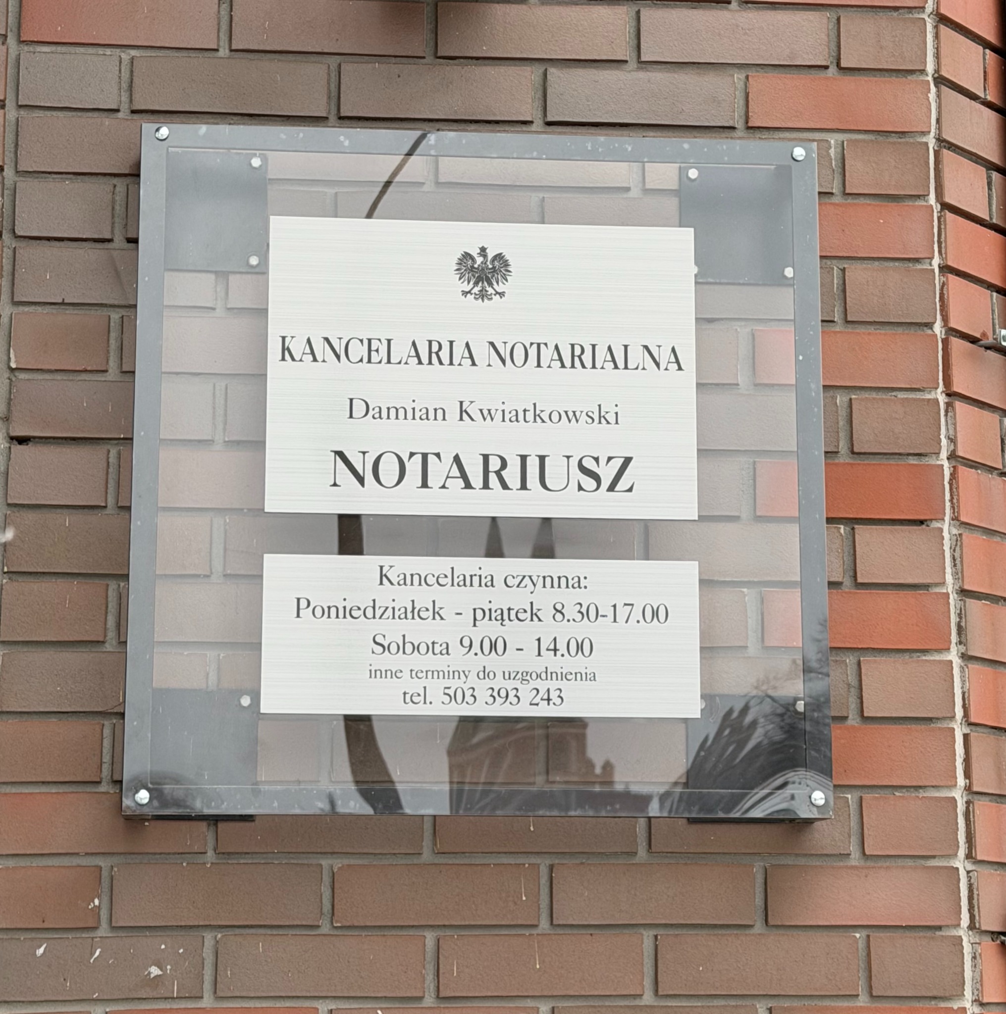 Tabliczka informacyjna na ceglanej ścianie z napisem 'Kancelaria Notarialna Damian Kwiatkowski Notariusz' oraz godzinami otwarcia.