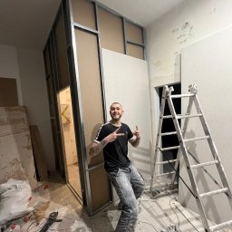 Dmytro Roi - Konstrukcja ścianki działowej z płyt karton-gips, widoczna metalowa rama i płyty. Na pierwszym planie uśmiechnięty mężczyzna w trakcie pracy, w tle drabina i narzędzia.