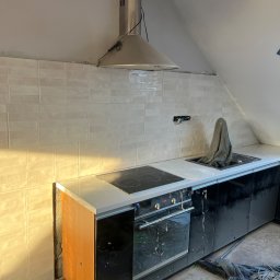 Dmytro Roi - Remont kuchni na poddaszu: białe, błyszczące płytki ułożone na ścianie, czarne szafki kuchenne z jasnym blatem, widoczny okap. W tle skos dachu i bałagan remontowy.