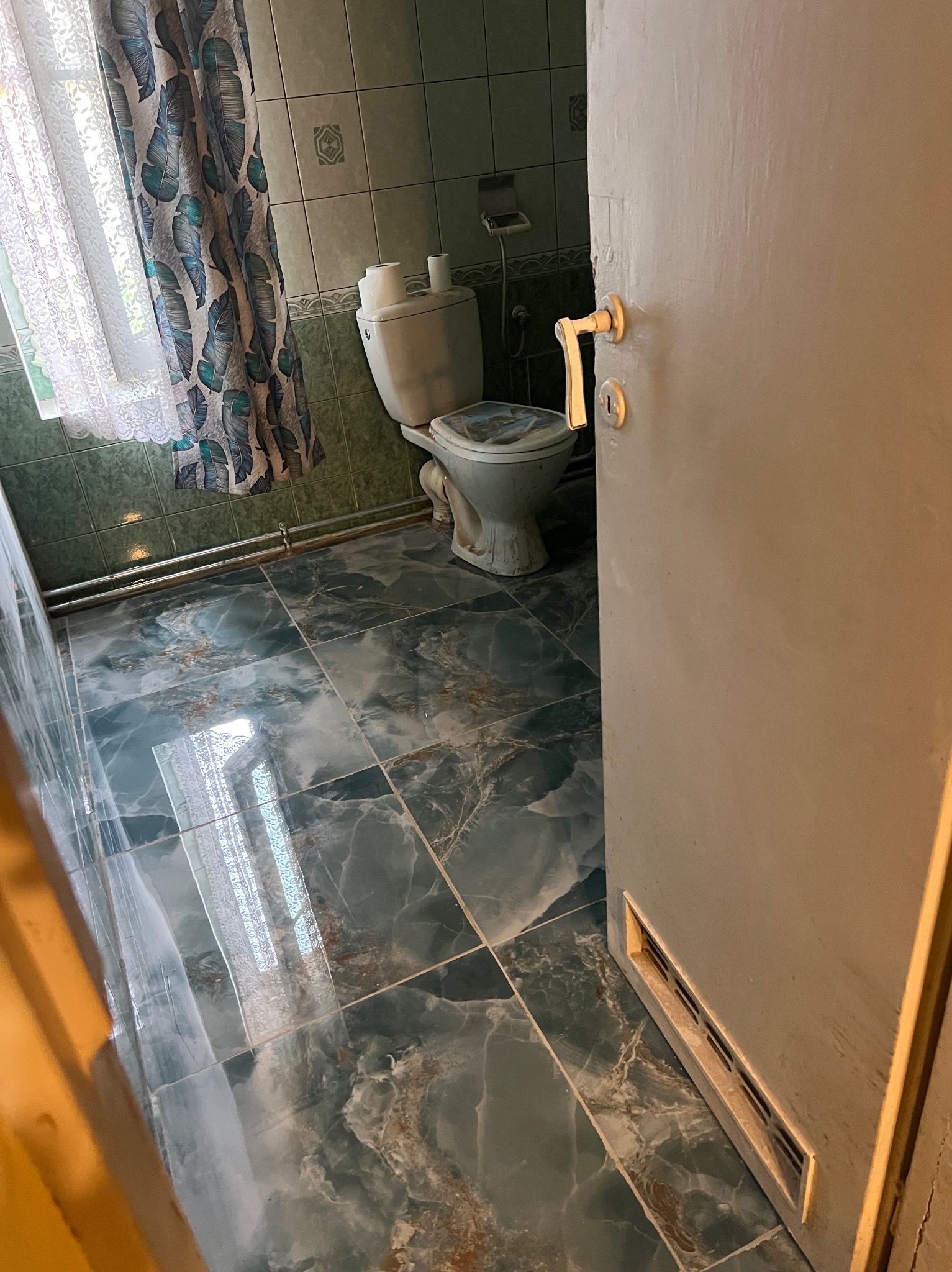 Ukończona łazienka z zielonymi płytkami podłogowymi i ściennymi, widoczna toaleta, okno z zasłoną i uchylone drzwi. Ujęcie z perspektywy wejścia.