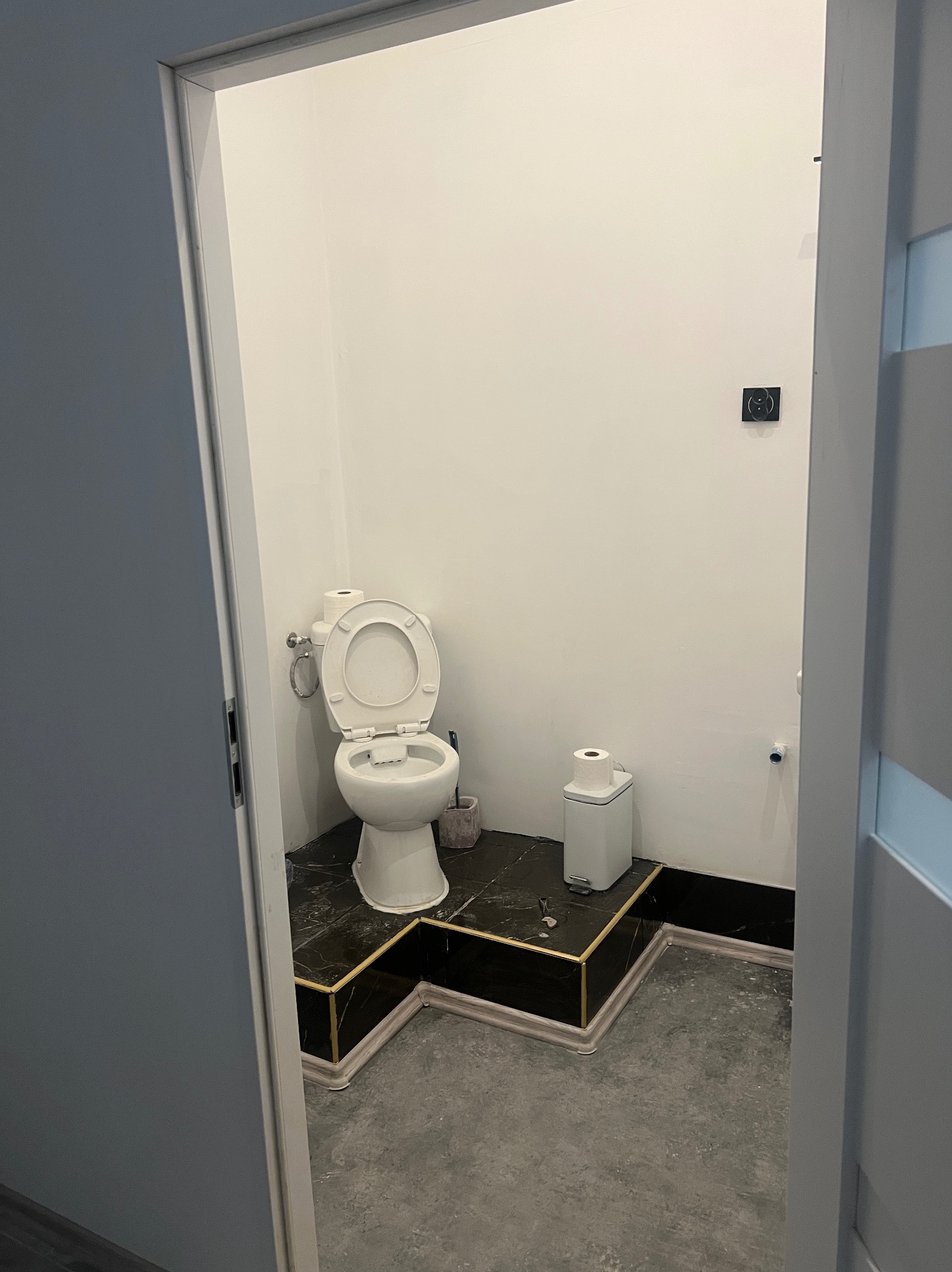 Wykończona łazienka z toaletą na podeście z czarnego marmuru ze złotymi akcentami, widoczna zza uchylonych drzwi. Minimalistyczny design, białe ściany i szara podłoga.