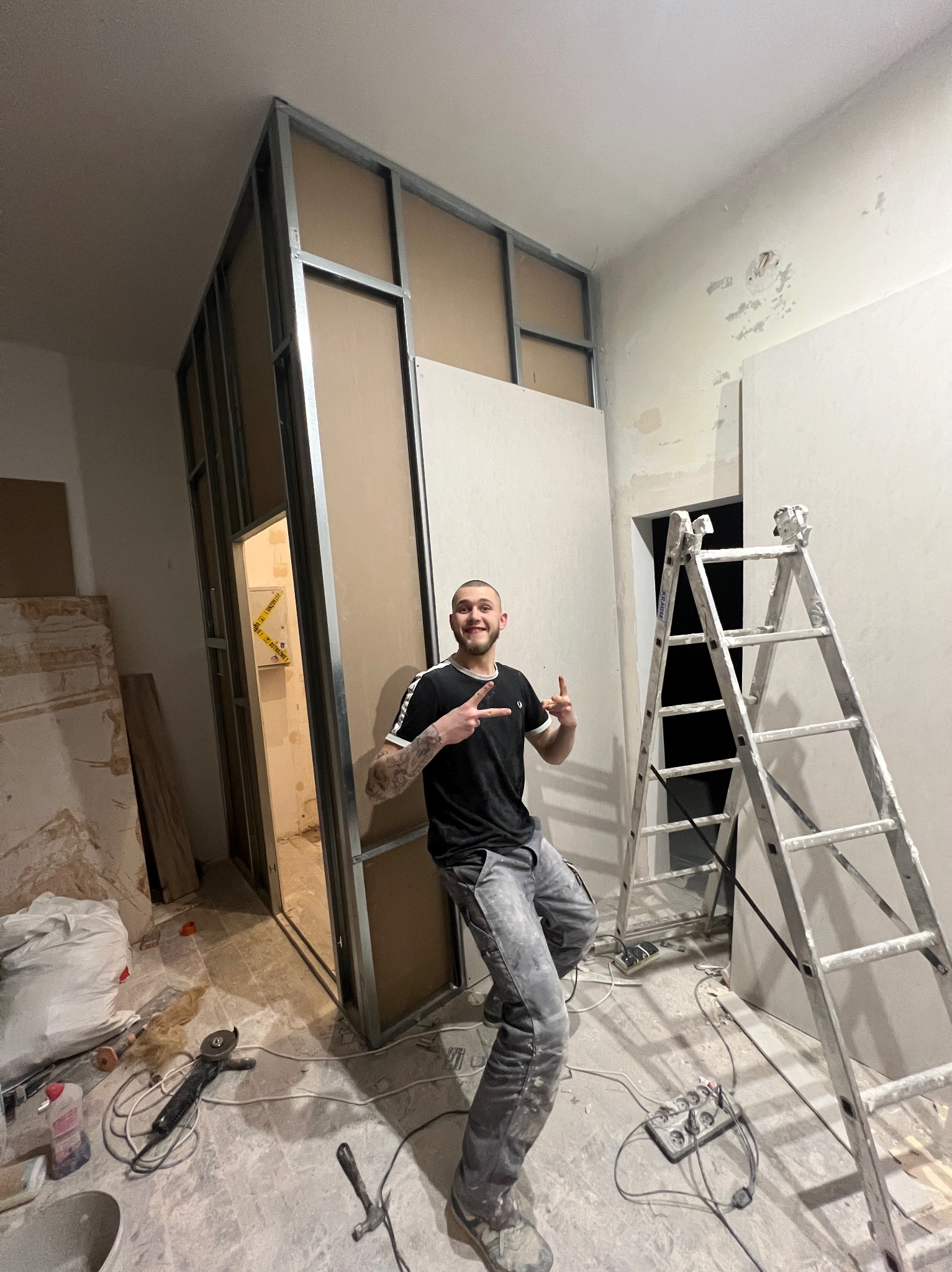 Konstrukcja ścianki działowej z płyt karton-gips, widoczna metalowa rama i płyty. Na pierwszym planie uśmiechnięty mężczyzna w trakcie pracy, w tle drabina i narzędzia.