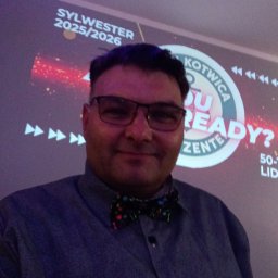 Zenon Kotwica - Mężczyzna w muszce na tle projekcji 'Sylwester 2025/2026 Kotwica'. Uśmiechnięty, w okularach, prezentuje się na tle imprezowej grafiki.