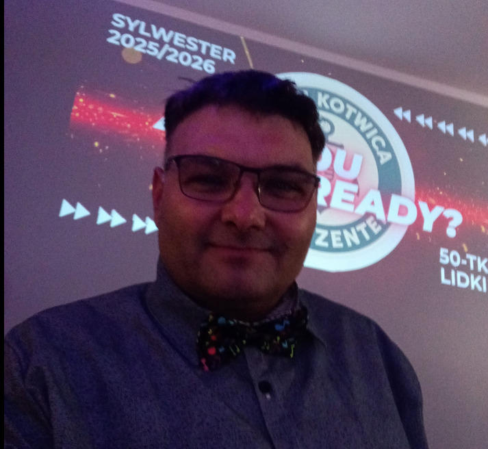 Mężczyzna w muszce na tle projekcji 'Sylwester 2025/2026 Kotwica'. Uśmiechnięty, w okularach, prezentuje się na tle imprezowej grafiki.