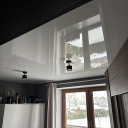 StretchDom-sufity napinane - Błyszczący, biały sufit napinany z odbiciem okna i zimowego krajobrazu. Widoczne czarne, okrągłe lampy sufitowe i fragment pokoju z szafą i oknem.