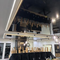 StretchDom-sufity napinane - Nowoczesne wnętrze z czarnym, lustrzanym sufitem podwieszanym i oświetleniem LED. Krzesła ustawione na stole, przygotowane do prac wykończeniowych.