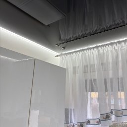 StretchDom-sufity napinane - Błyszcząca zabudowa kuchenna z białymi frontami, oświetleniem LED podszafkowym i delikatnymi firankami w oknie. Widoczny fragment okna z widokiem na zewnątrz.