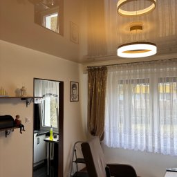 StretchDom-sufity napinane - Błyszczący sufit napinany w salonie z nowoczesnym oświetleniem. Widoczne okno z firankami, fotel i dekoracje na półkach. Refleksy światła podkreślają gładką powierzchnię sufitu.