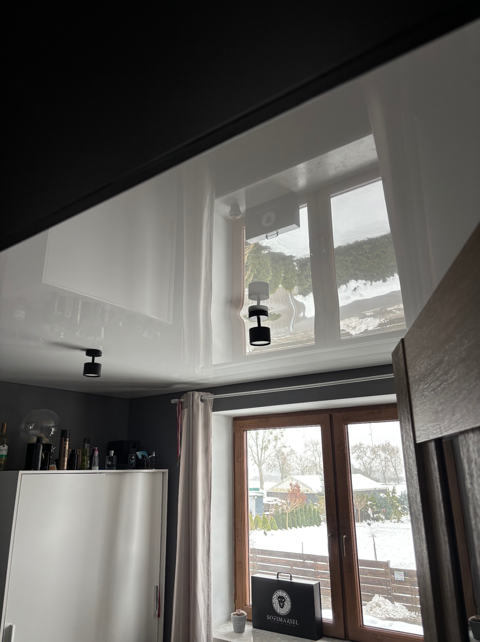 Błyszczący, biały sufit napinany z odbiciem okna i zimowego krajobrazu. Widoczne czarne, okrągłe lampy sufitowe i fragment pokoju z szafą i oknem.
