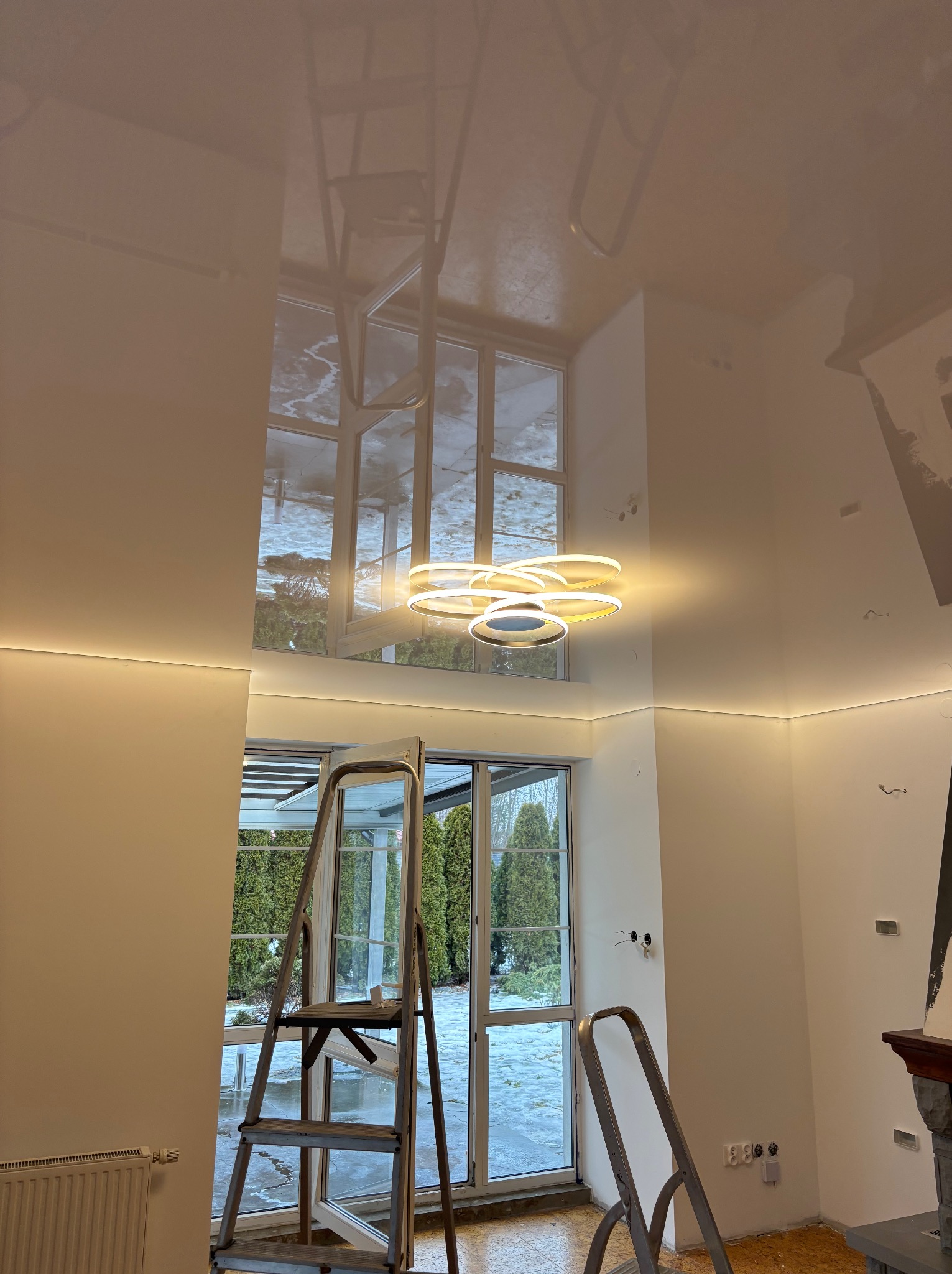 Białe wnętrze z wysokim połyskiem sufitu, nowoczesną lampą i oknami. Widoczne drabiny sugerują prace wykończeniowe w toku, z elementami instalacji oświetleniowej.