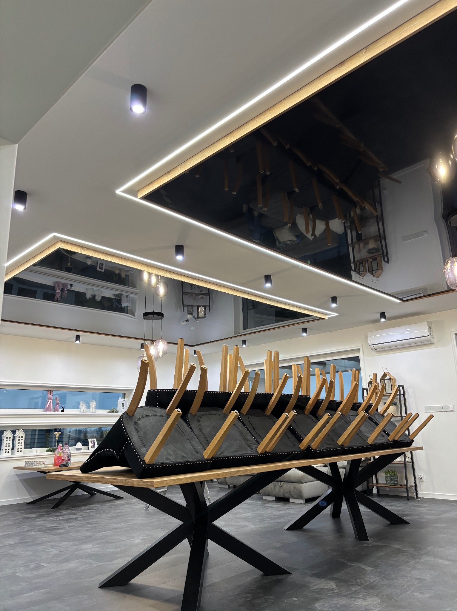 Nowoczesne wnętrze z czarnym, lustrzanym sufitem podwieszanym i oświetleniem LED. Krzesła odwrócone na stole w przestronnym pokoju z szarą podłogą.