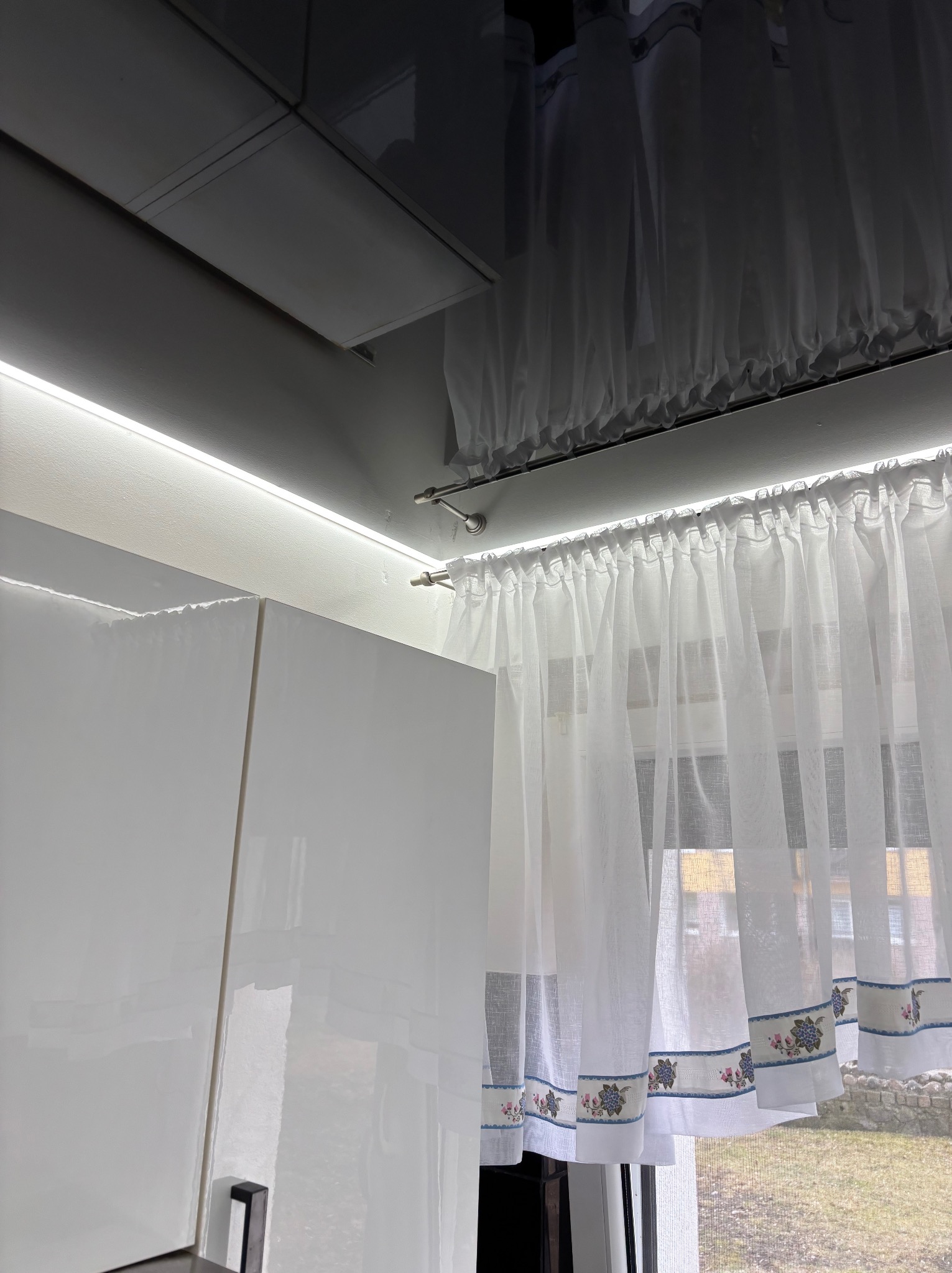 Błyszcząca zabudowa kuchenna z białymi frontami, oświetleniem LED podszafkowym i delikatnymi firankami w oknie. Widoczny fragment okna z widokiem na zewnątrz.