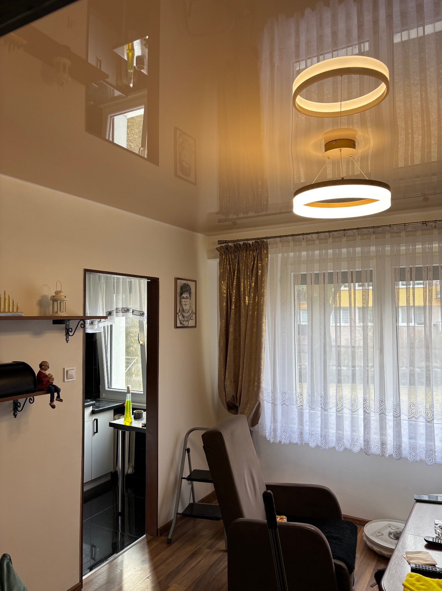 Błyszczący sufit napinany w salonie z nowoczesnym oświetleniem. Widoczne okno z firankami, fotel i dekoracje na półkach. Refleksy światła podkreślają gładką powierzchnię sufitu.