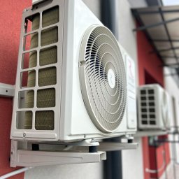 AIR-CTIC MARCIN LUBACZEWSKI - Zamontowana, biała jednostka zewnętrzna klimatyzacji na ścianie budynku z elementami czerwieni. Widoczna druga jednostka w tle, pod zadaszeniem.