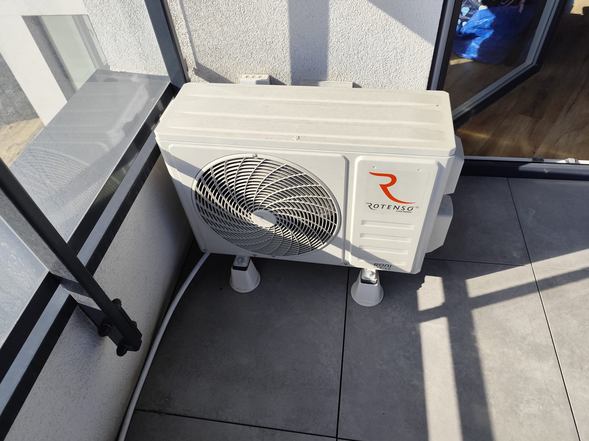 Biała jednostka zewnętrzna klimatyzacji Rotenso na balkonie z szarymi płytkami, widoczna z boku, z fragmentem okna i ściany w tle. Jasne, naturalne światło.
